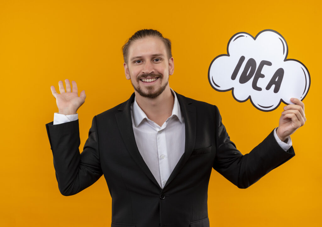 handsome-business-man-wearing-suit-holding-word-idea-speech-bubble-looking-camera-happy-positive-smiling-cheerfully-standing-orange-background